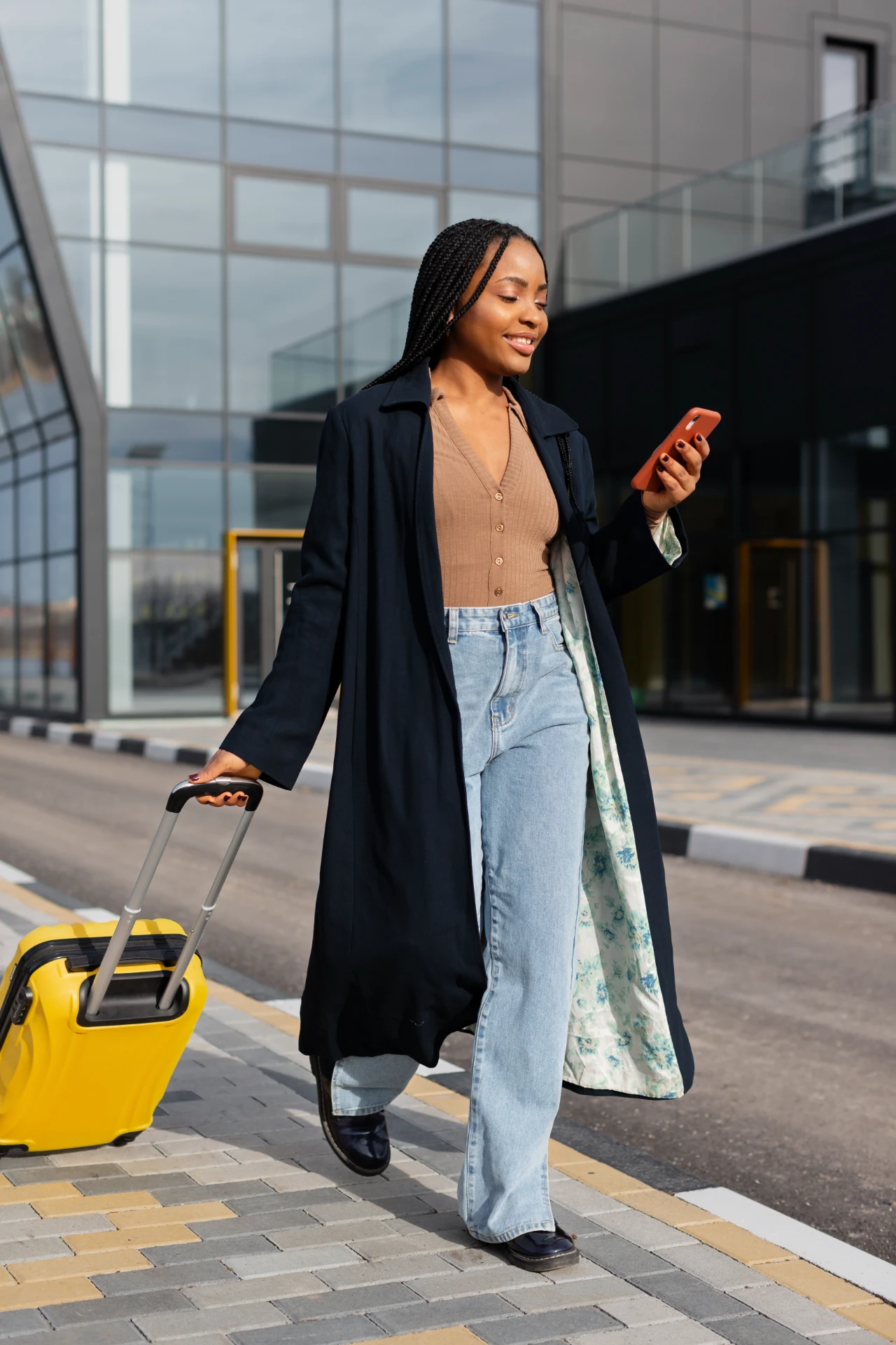 Traveler walking outside an airport terminal with a yellow suitcase and a smartphone